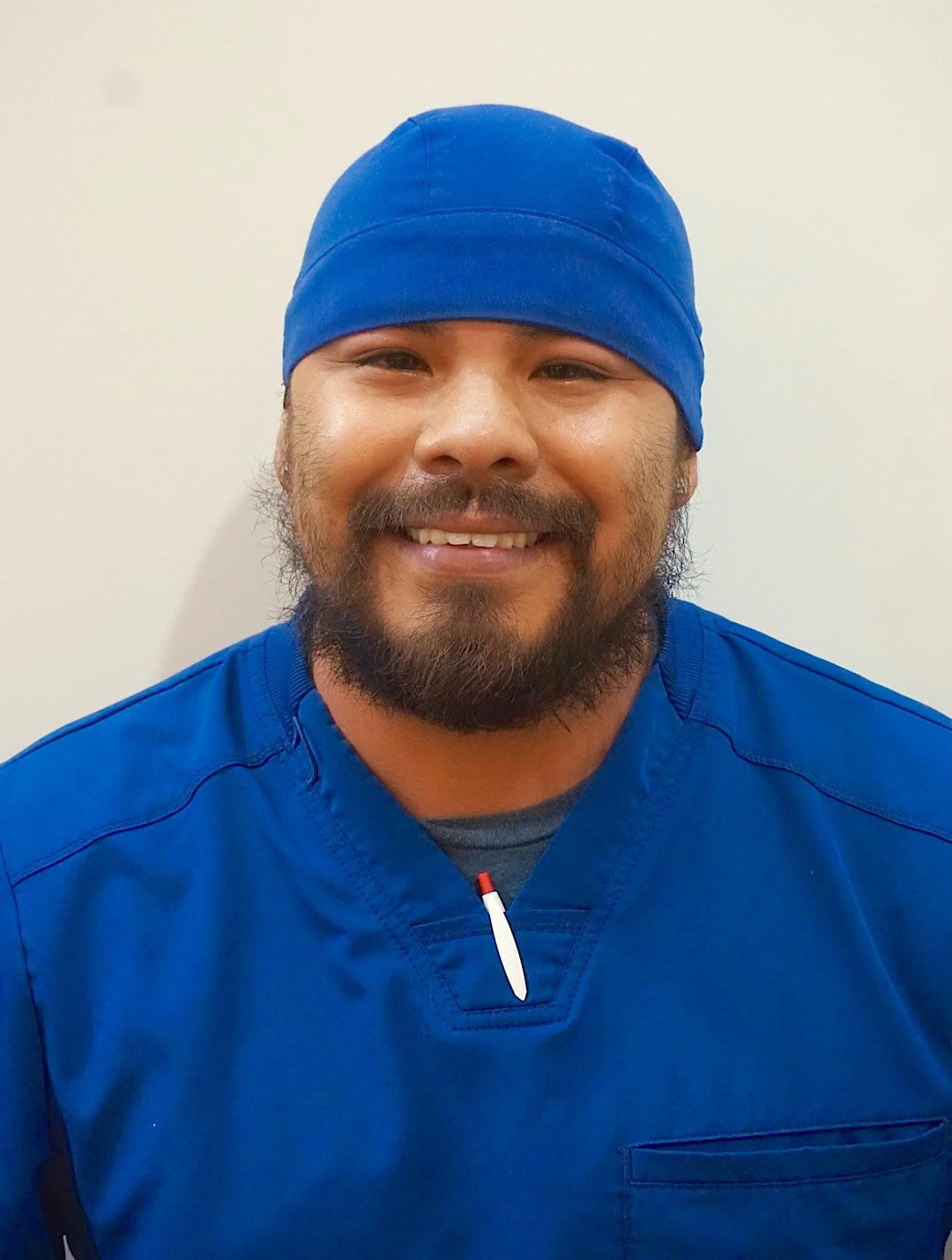 The image shows a man wearing a blue surgical gown and cap, smiling at the camera with his head tilted slightly downward.