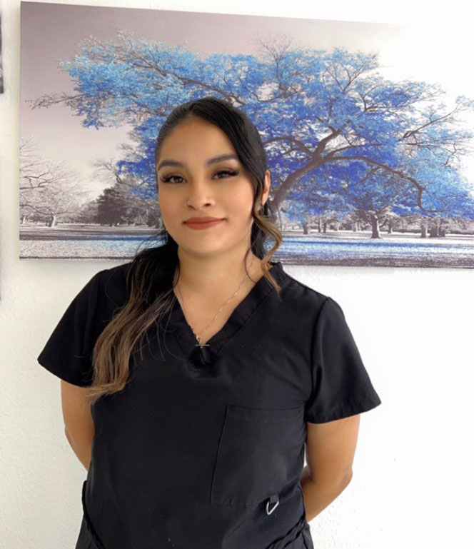 The image shows a woman standing in front of a framed picture, wearing scrubs and smiling at the camera.