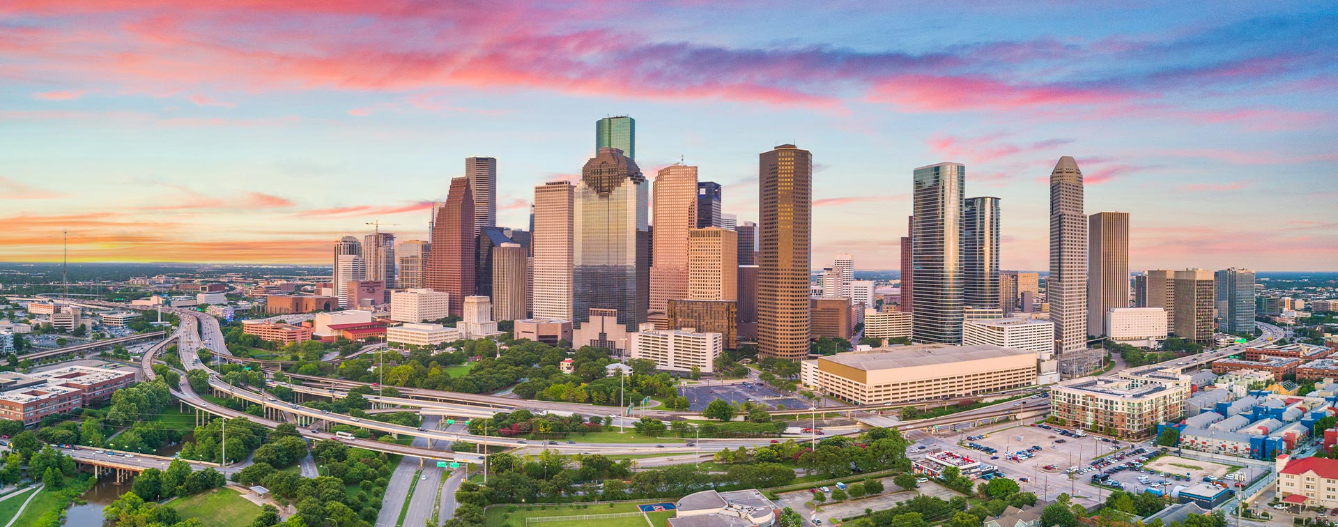 The image displays a panoramic view of a city skyline at sunset, with a prominent downtown area featuring multiple high-rise buildings under a vibrant sky with shades of pink and orange, suggesting a peaceful evening in an urban setting.