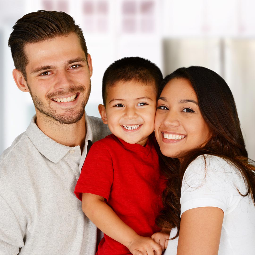 A family posing together with smiles, featuring an adult couple holding a child, set against a domestic backdrop.