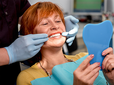 The image shows a woman sitting in a dental chair with an open mouth, holding a blue toothbrush, while a dentist works on her teeth.
