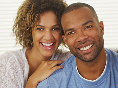 The image shows a man and a woman posing together with smiles, the man has short hair and a beard, and they appear to be standing in front of a light-colored background.