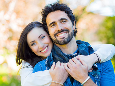 The image depicts two people embracing each other outdoors  they appear happy and are smiling at the camera.