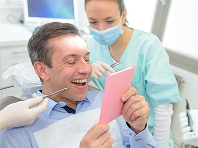 The image features a person sitting in a dental chair with a smile on their face, looking at a pink card held by another individual who appears to be a dentist. They are surrounded by medical equipment typically found in a dental office, and there is a third individual present who seems to be observing the situation.