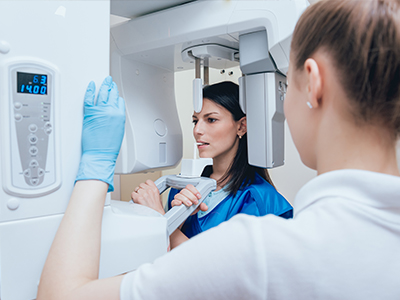 A woman wearing a blue jacket stands next to a large, modern CT scanner machine, with another person partially visible behind her, examining their face through the scanner s window.