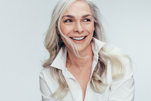 The image features a woman with gray hair smiling at the camera  she s wearing a white top and has her hair styled short, and she appears to be indoors with a neutral background.
