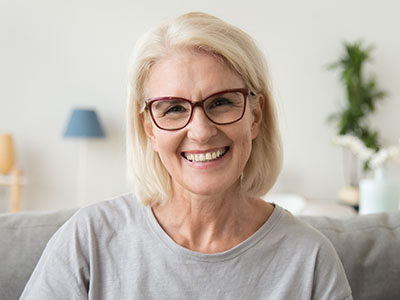 A woman with short blonde hair wearing glasses, smiling at the camera, sitting on a couch indoors.