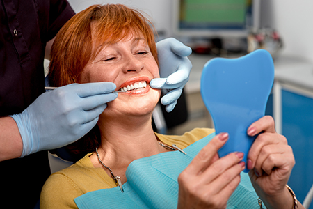 A woman sitting in a dental chair with a blue face mask on her face, holding up a tooth model while smiling at the camera.