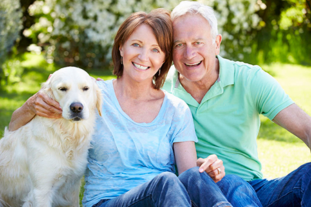 The image shows an older couple sitting on grass with a dog between them, smiling towards the camera.