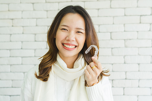 A woman with a smile holds a white object up to her face, standing against a brick wall background.