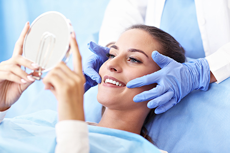 Woman with blue gloves holding up mirror to show face while seated on dental chair.