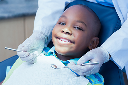 A young boy is seated in a dental chair with his mouth open, receiving dental care from a dentist who is smiling at him.