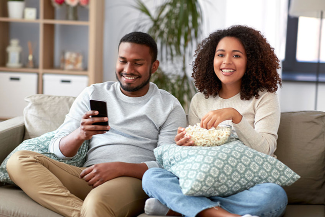 A man and woman sitting on a couch, sharing a moment while enjoying popcorn, with the man smiling at the camera and the woman looking down at her phone.