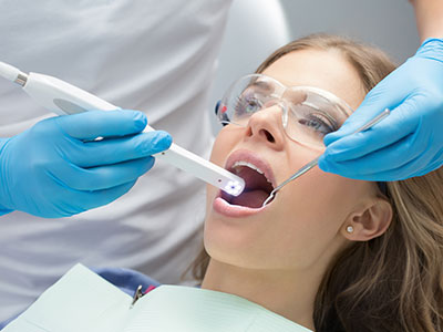 Woman wearing blue gloves and holding a toothbrush-like device over a woman's mouth during dental procedure.