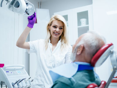 A woman wearing a white lab coat stands beside an older man in a medical setting, both are smiling, with the woman pointing at something above the man's head.
