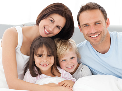 The image shows a family of four people—a man, a woman, and two children—smiling and posing together on a bed.