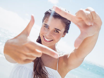A woman with long hair is holding up two fingers at her eye level, smiling, with a bright background and beach setting.